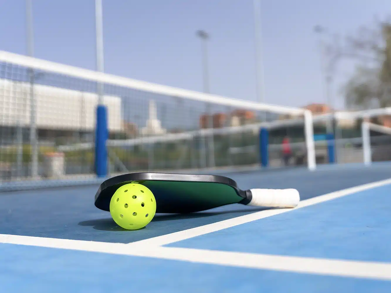 Pickleball paddle and ball resting on a blue outdoor pickleball court.